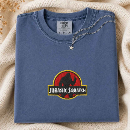 A folded blue t-shirt with a 'Jurassic Squatch' parody graphic on the front, styled on a cream knit blanket with a silver star necklace.