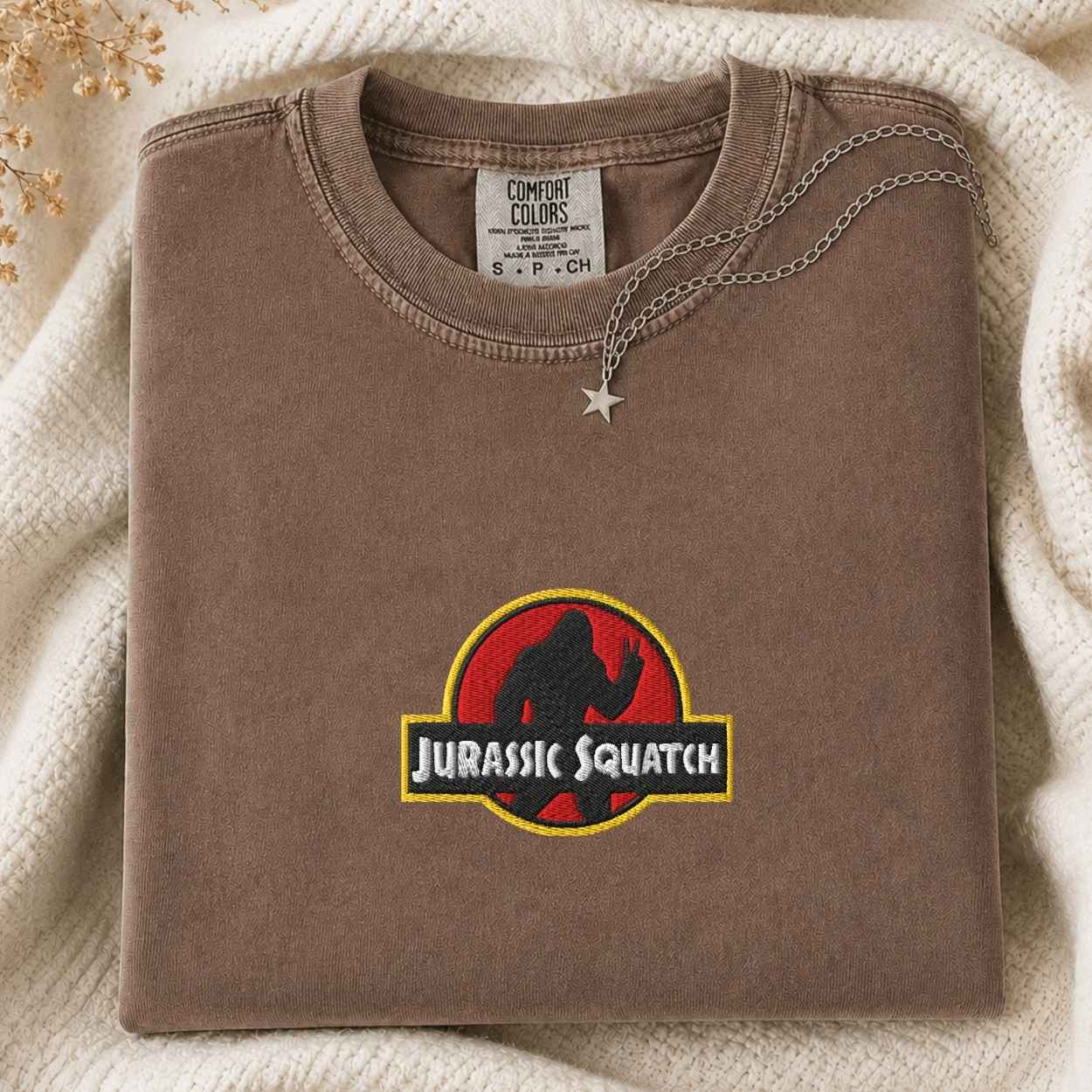 A folded brown Comfort Colors t-shirt with a 'Jurassic Squatch' graphic on the front, styled on a cream knit blanket with a silver star necklace.