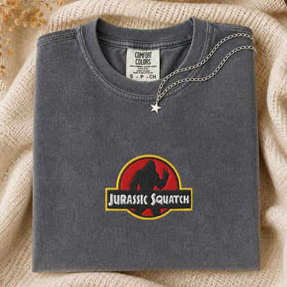 A folded dark gray t-shirt featuring a 'Jurassic Squatch' graphic on the front, styled on a beige knit blanket with a silver star necklace draped over the collar.
