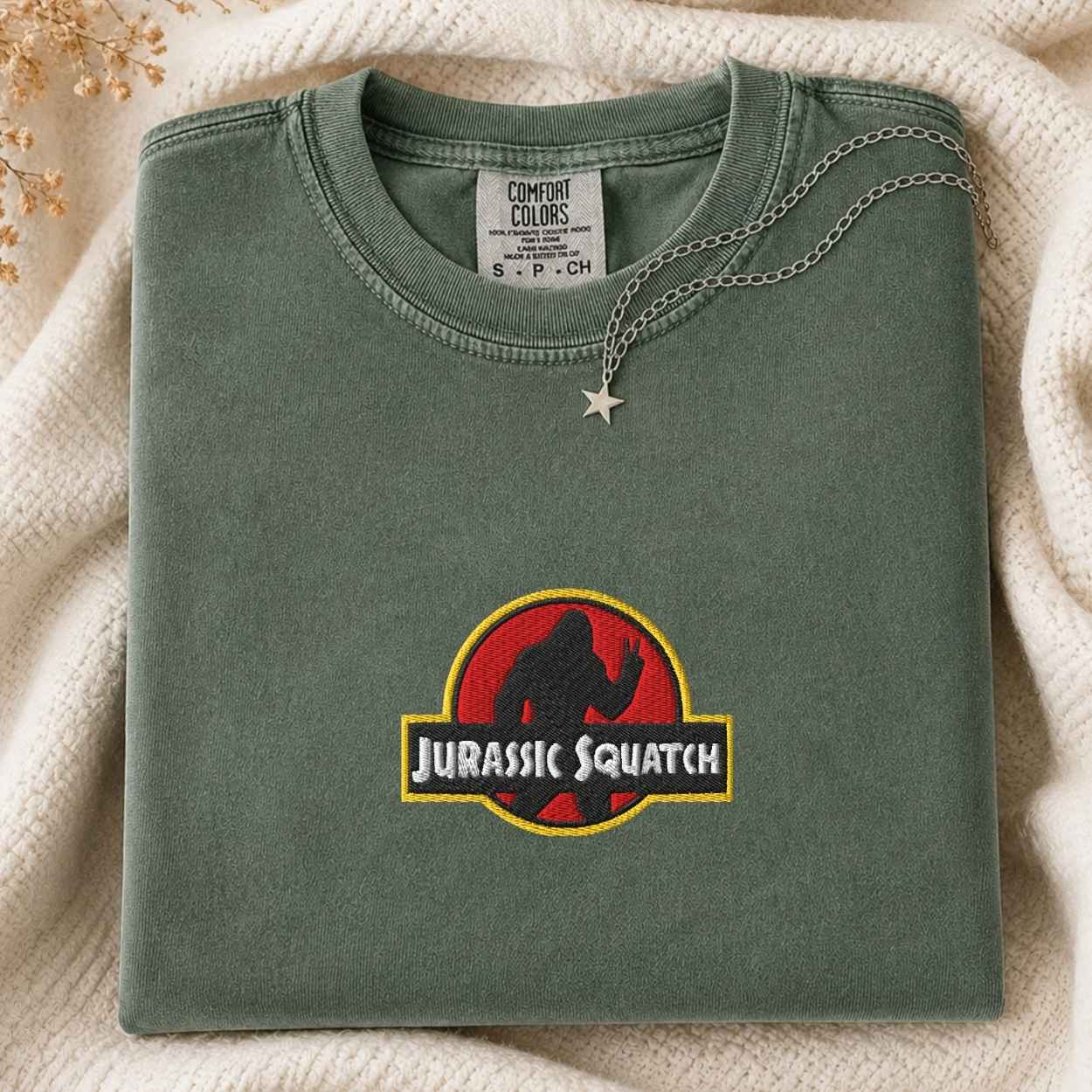 A folded sage green Comfort Colors t-shirt featuring a 'Jurassic Squatch' graphic on the front, styled on a cream knit blanket with a silver necklace.