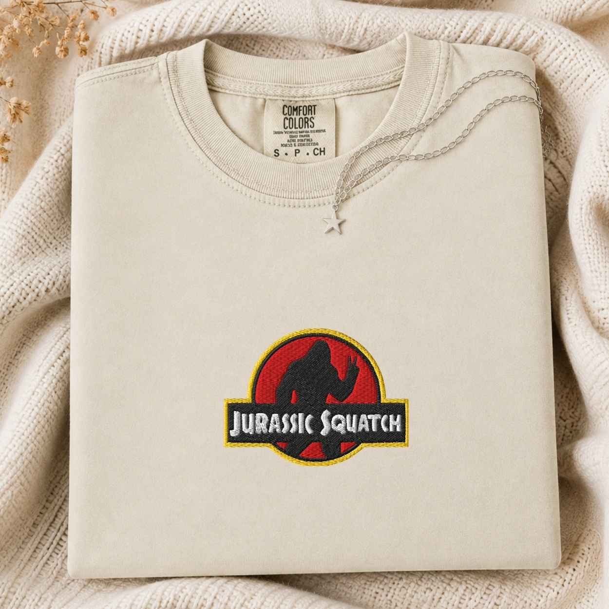 A folded cream-colored sweatshirt with a 'Jurassic Squatch' parody graphic on the front, styled on a cozy cream knit blanket with dried flowers.