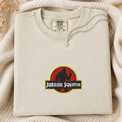 A folded cream-colored sweatshirt with a 'Jurassic Squatch' parody graphic on the front, styled on a cozy cream knit blanket with dried flowers.