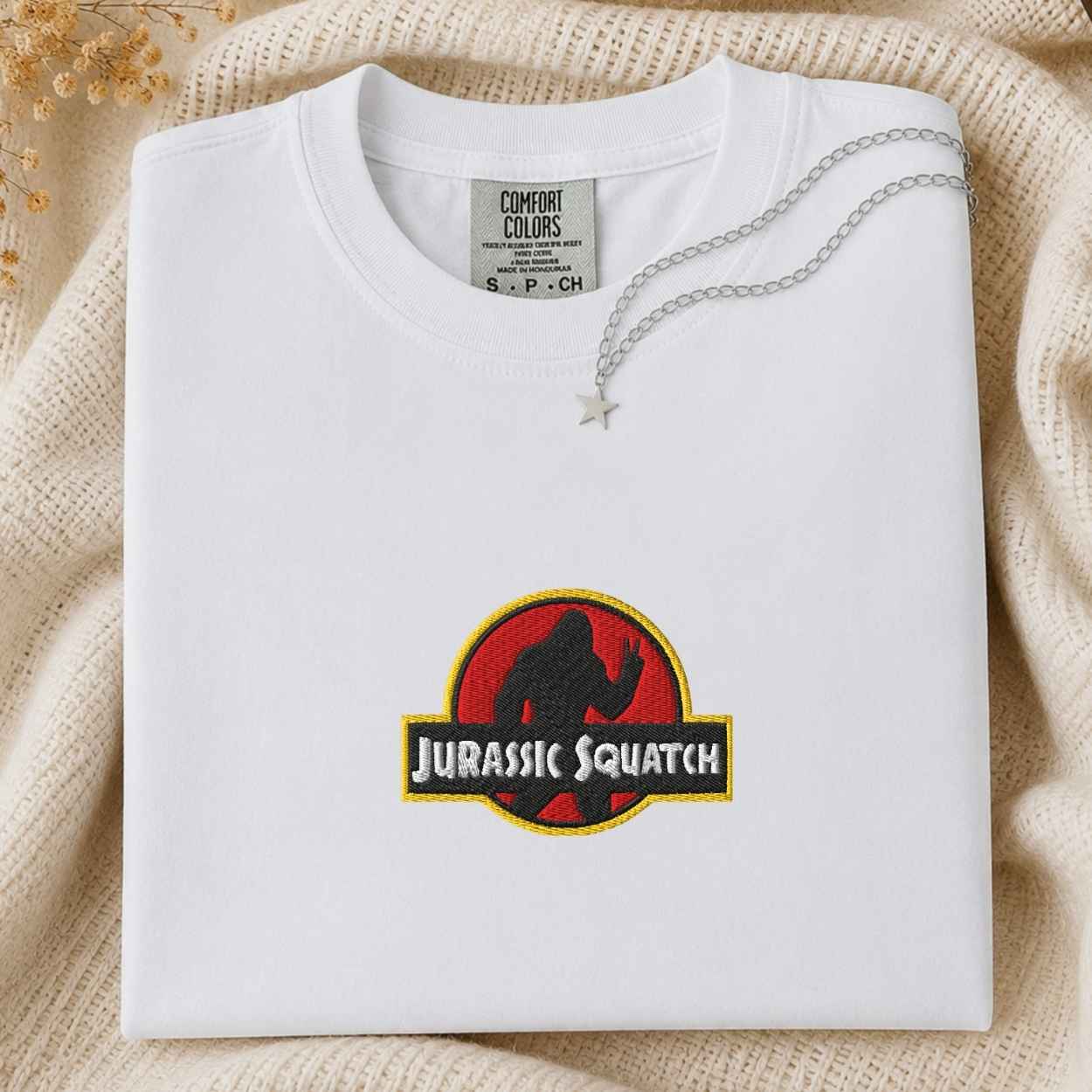 A folded white Comfort Colors t-shirt featuring a custom 'Jurassic Squatch' parody graphic on the front, styled on a beige knit blanket with a silver necklace.
