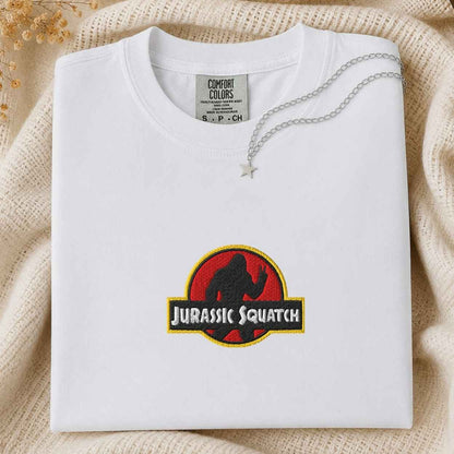 A folded white Comfort Colors t-shirt featuring a custom 'Jurassic Squatch' parody graphic on the front, styled on a beige knit blanket with a silver necklace.