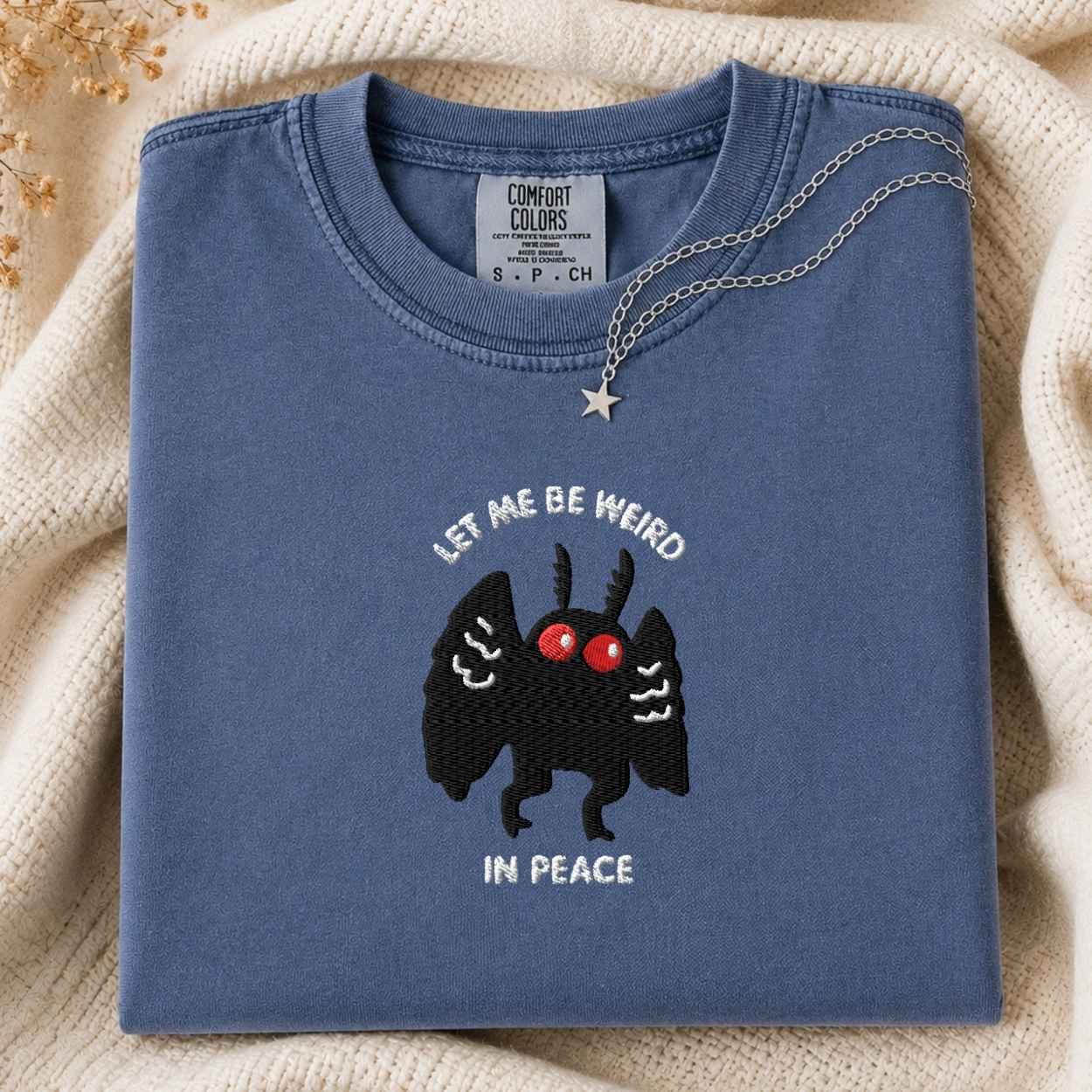A folded slate blue Comfort Colors t-shirt with a black embroidered mothman character, red eyes, and the text 'LET ME BE WEIRD IN PEACE'. A silver necklace with a star pendant is draped over the collar.