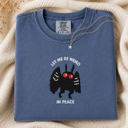 A folded slate blue Comfort Colors t-shirt with a black embroidered mothman character, red eyes, and the text 'LET ME BE WEIRD IN PEACE'. A silver necklace with a star pendant is draped over the collar.