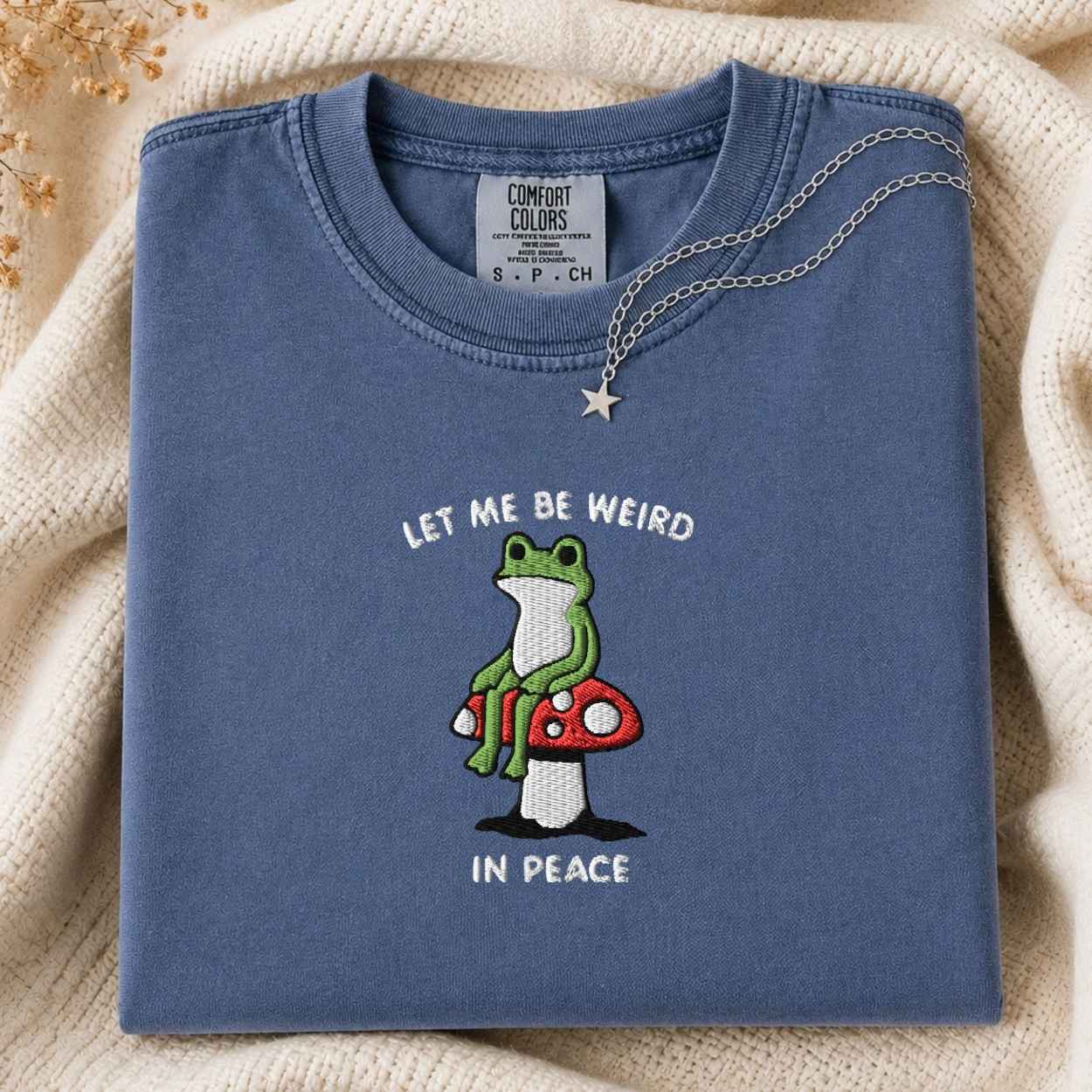 A folded slate blue t-shirt with a graphic of a green frog sitting on a red and white mushroom, with the text 'LET ME BE WEIRD IN PEACE'. The t-shirt is styled on a cream-colored knit blanket with a silver necklace draped over the collar.
