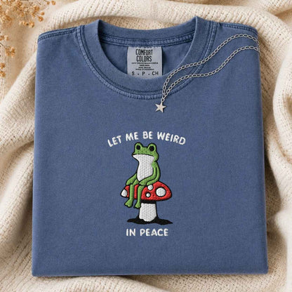 A folded slate blue t-shirt with a graphic of a green frog sitting on a red and white mushroom, with the text 'LET ME BE WEIRD IN PEACE'. The t-shirt is styled on a cream-colored knit blanket with a silver necklace draped over the collar.