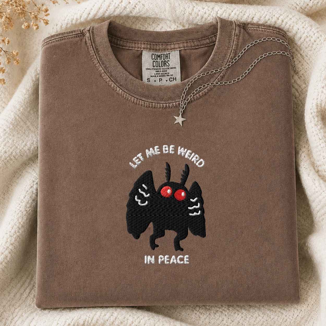 A folded brown tshirt with a graphic of a black mothman with red eyes and the text 'LET ME BE WEIRD IN PEACE' on the front, styled on a cream knit blanket with a silver necklace.