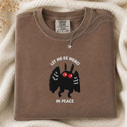 A folded brown tshirt with a graphic of a black mothman with red eyes and the text 'LET ME BE WEIRD IN PEACE' on the front, styled on a cream knit blanket with a silver necklace.