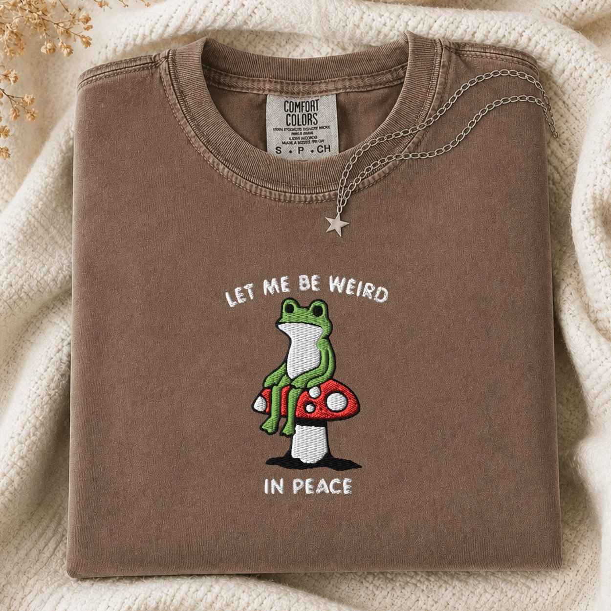 A folded brown crewneck sweatshirt with a graphic of a green frog sitting on a red mushroom and the text 'LET ME BE WEIRD IN PEACE'. A silver star pendant necklace is draped over the collar.