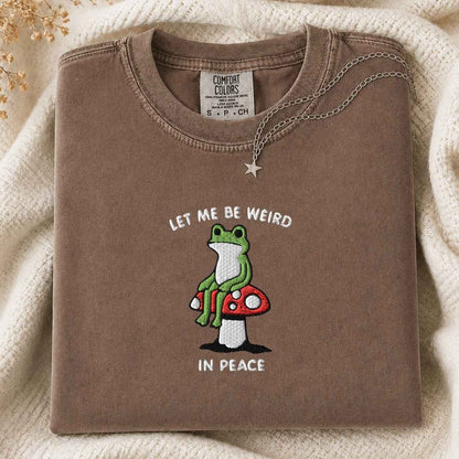 A folded brown crewneck sweatshirt with a graphic of a green frog sitting on a red mushroom and the text 'LET ME BE WEIRD IN PEACE'. A silver star pendant necklace is draped over the collar.