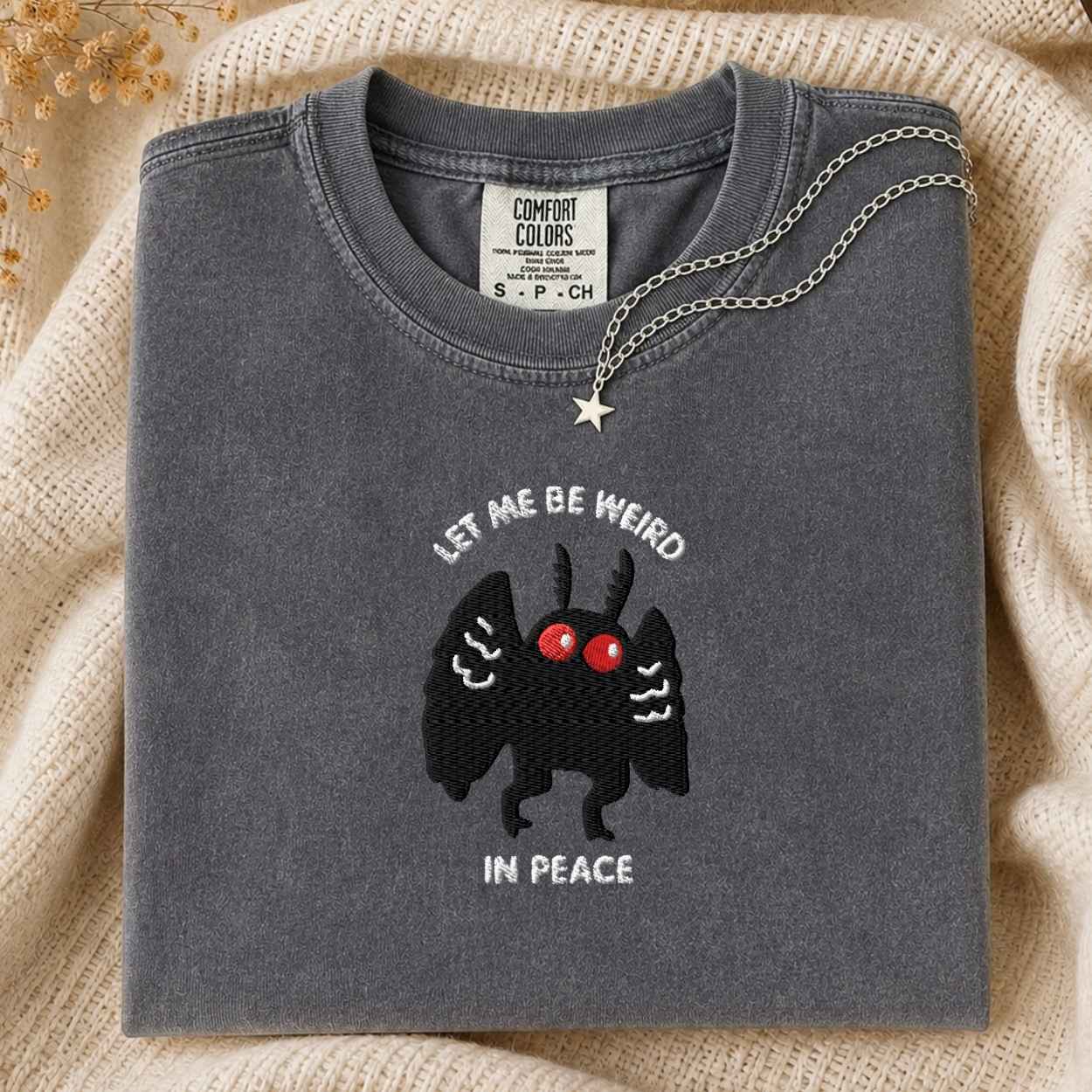 A folded dark gray t-shirt with a black winged creature graphic and the text 'LET ME BE WEIRD IN PEACE' on the front, styled on a beige knit blanket with a silver necklace draped over the collar.