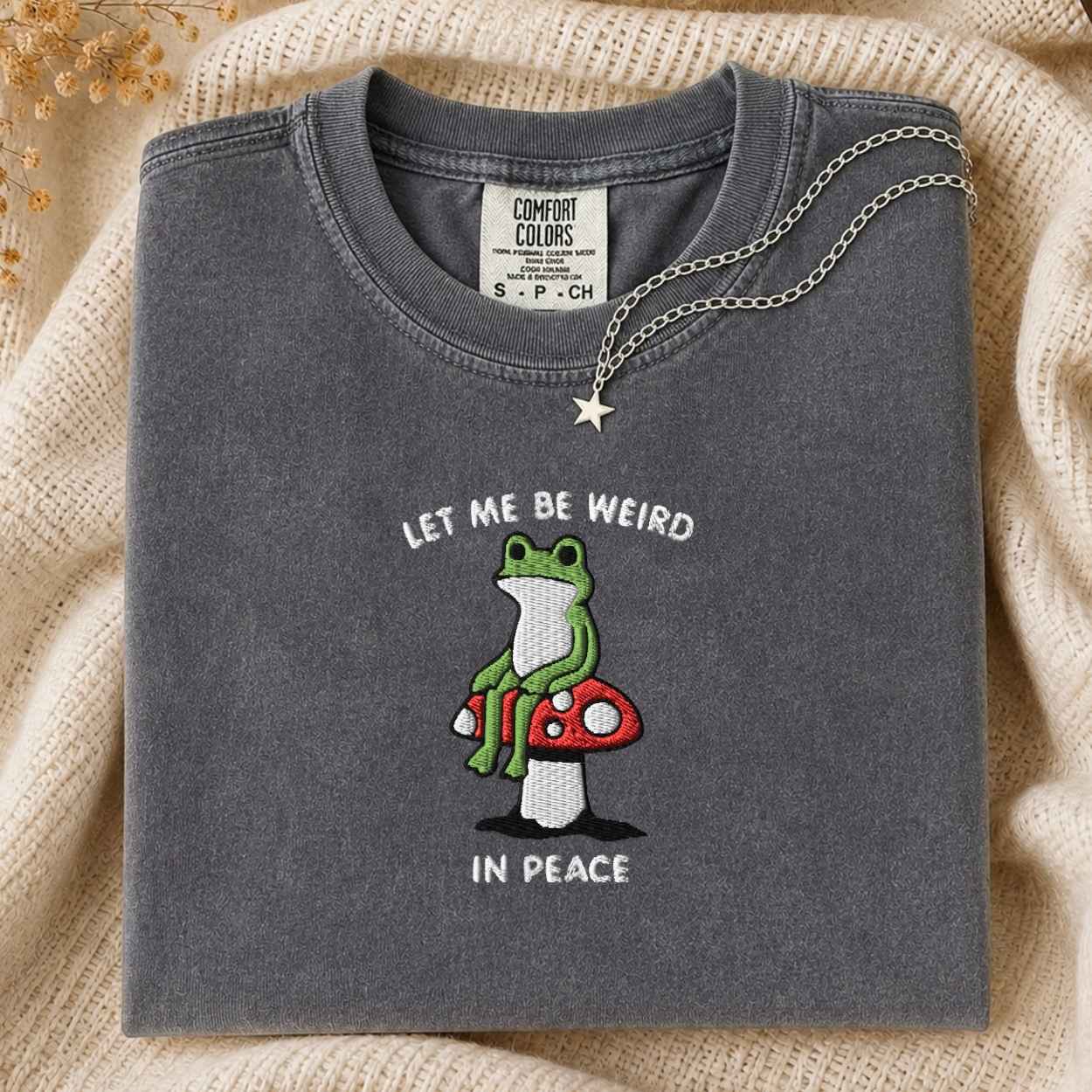 A folded dark gray t-shirt with a graphic of a green frog sitting on a red mushroom, with the text 'LET ME BE WEIRD IN PEACE'. A silver necklace with a star pendant is draped over the collar.