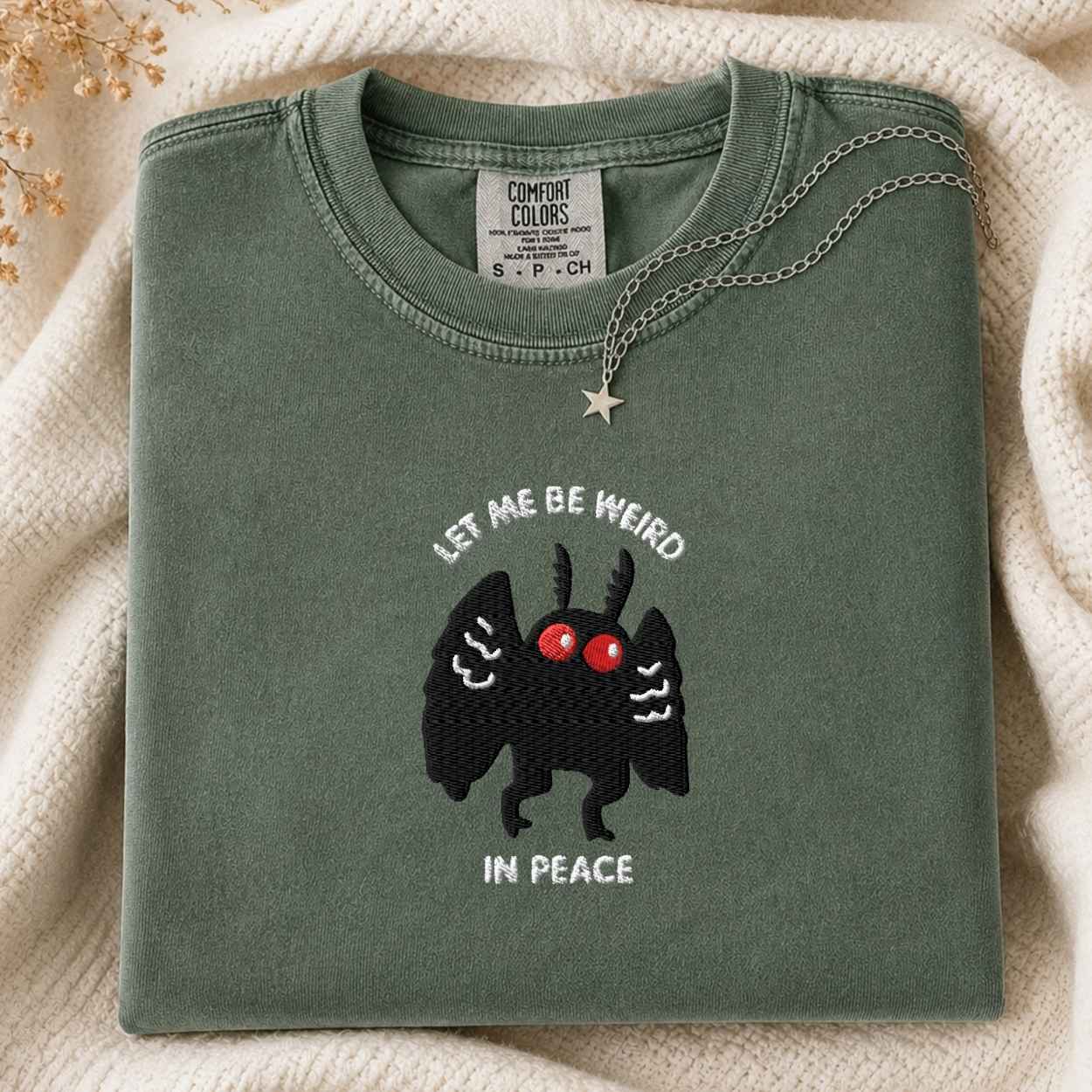 A folded sage green t-shirt with a graphic of a black mothman with red eyes and the text 'LET ME BE WEIRD IN PEACE'. A silver necklace with a star pendant is draped over the collar.