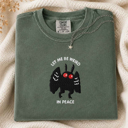 A folded sage green t-shirt with a graphic of a black mothman with red eyes and the text 'LET ME BE WEIRD IN PEACE'. A silver necklace with a star pendant is draped over the collar.