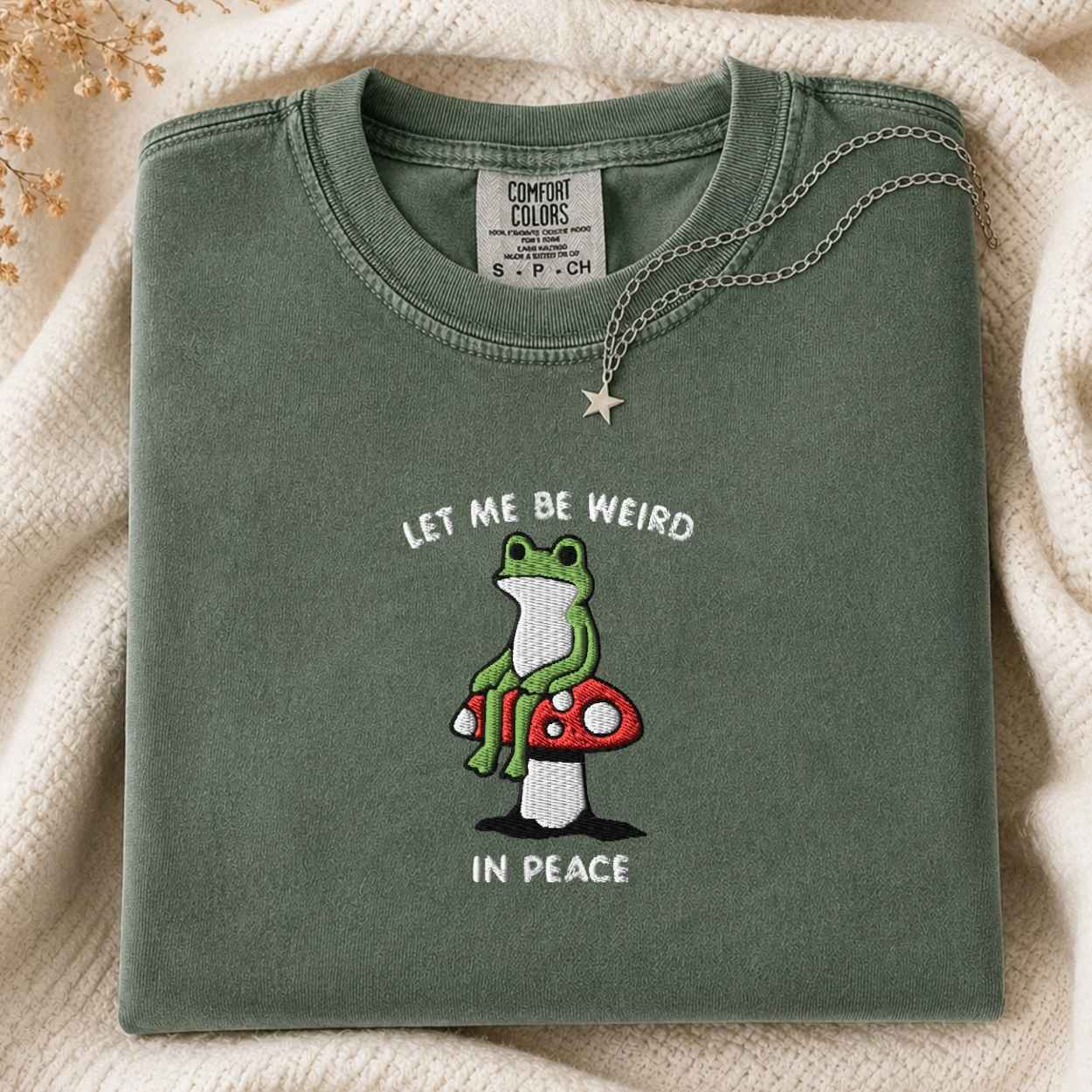 A folded sage green t-shirt with a graphic of a green frog sitting on a red and white mushroom, with the text 'LET ME BE WEIRD IN PEACE'. A silver star necklace is draped over the collar.