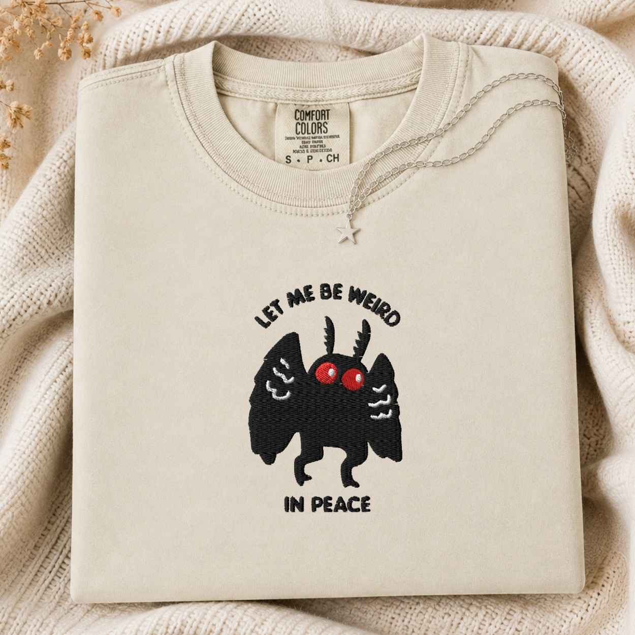 A folded cream sweatshirt with a graphic of a black winged creature with red eyes and the text 'LET ME BE WEIRD IN PEACE' on the front, styled on a textured cream knit blanket.