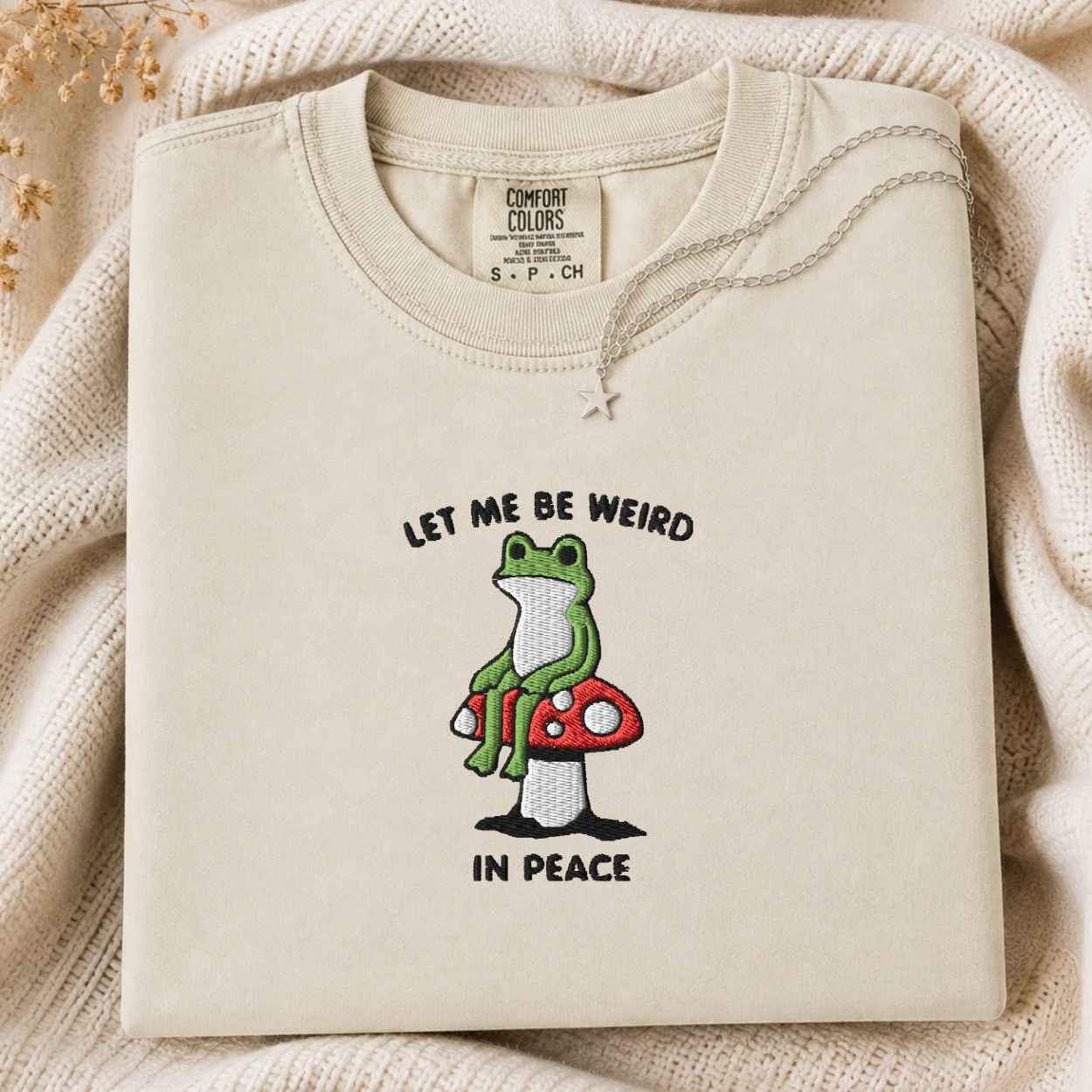 A folded cream-colored tshirt with a graphic of a green frog sitting on a red mushroom, accompanied by the text 'LET ME BE WEIRD IN PEACE'. The tshirt is displayed on a cozy, textured cream knit blanket with dried flowers in the corner.