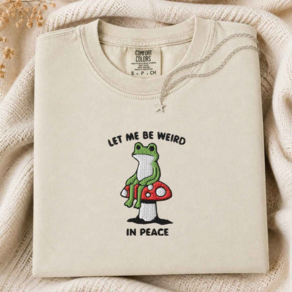 A folded cream-colored tshirt with a graphic of a green frog sitting on a red mushroom, accompanied by the text 'LET ME BE WEIRD IN PEACE'. The tshirt is displayed on a cozy, textured cream knit blanket with dried flowers in the corner.