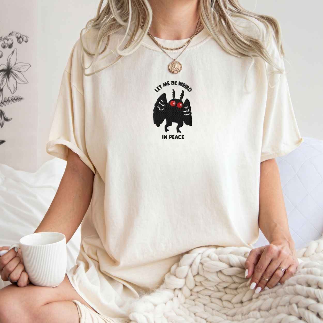 A woman wearing an oversized cream-colored t-shirt with a black and red graphic of a winged creature and the text 'LET ME BE WEIRD IN PEACE'. She is sitting in a cozy indoor setting with a white mug and a chunky knit blanket.