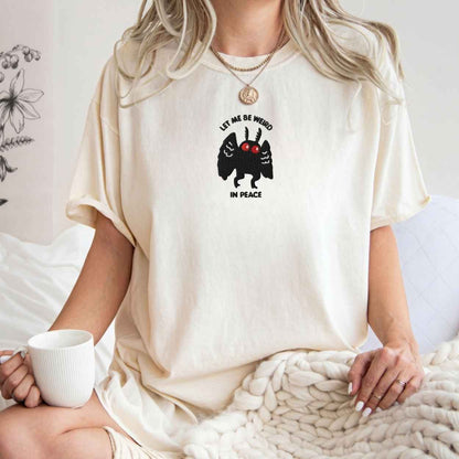 A woman wearing an oversized cream-colored t-shirt with a black and red graphic of a winged creature and the text 'LET ME BE WEIRD IN PEACE'. She is sitting in a cozy indoor setting with a white mug and a chunky knit blanket.