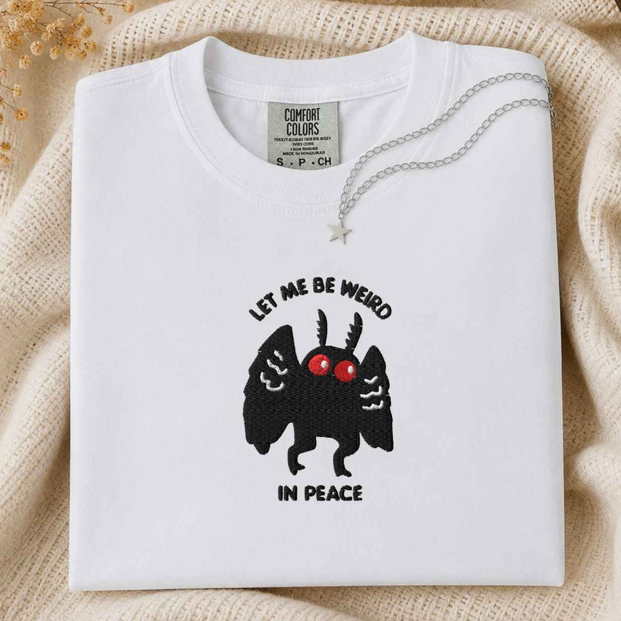 A folded white Comfort Colors t-shirt with a black cartoon mothman  graphic and the text 'LET ME BE WEIRD IN PEACE' on the front, styled on a beige knit blanket with a silver necklace.