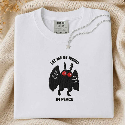 A folded white Comfort Colors t-shirt with a black cartoon mothman  graphic and the text 'LET ME BE WEIRD IN PEACE' on the front, styled on a beige knit blanket with a silver necklace.