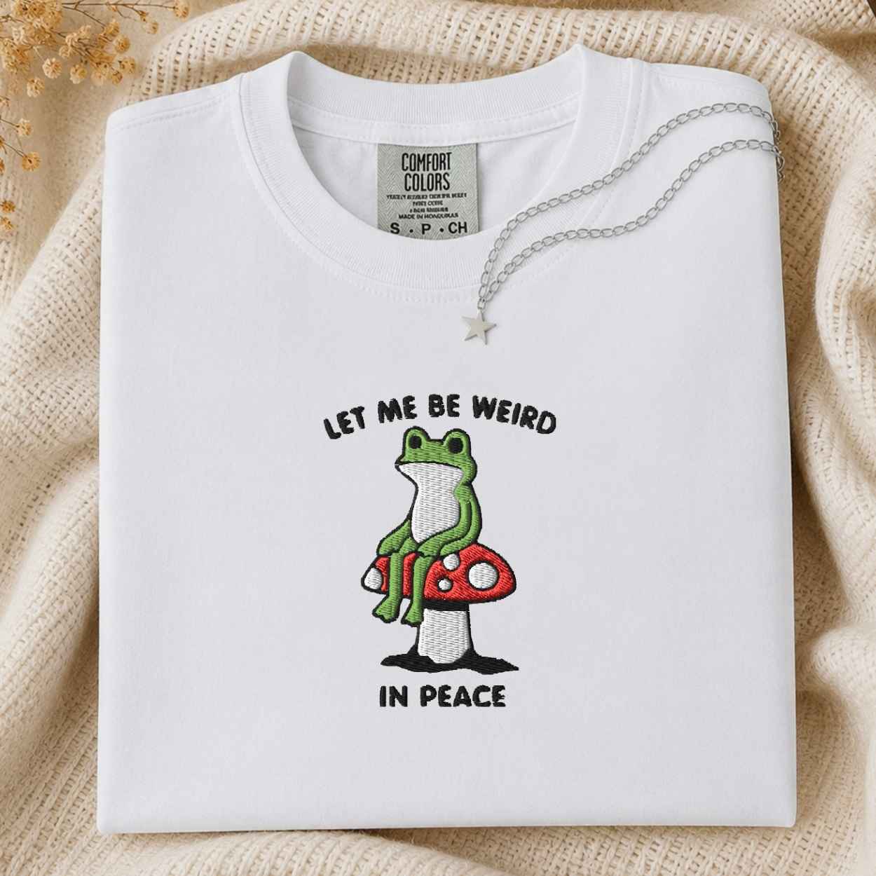 A folded white Comfort Colors t-shirt with a graphic of a green frog sitting on a red mushroom and the text 'LET ME BE WEIRD IN PEACE'. A silver necklace with a star pendant is draped over the collar.