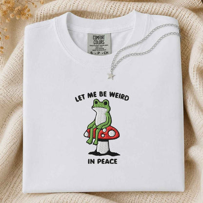 A folded white Comfort Colors t-shirt with a graphic of a green frog sitting on a red mushroom and the text 'LET ME BE WEIRD IN PEACE'. A silver necklace with a star pendant is draped over the collar.