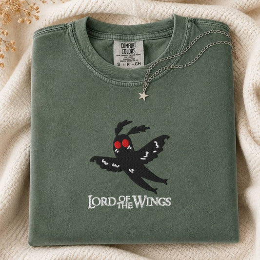 A folded sage green Comfort Colors t-shirt featuring a black mothman graphic with red eyes and the text 'LORD OF THE WINGS' embroidered below it. The shirt is styled on a cream knit blanket with a silver star necklace draped over the collar.