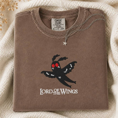 A folded brown sweatshirt with a black moth graphic and the words 'LORD OF THE WINGS' on the front, styled on a cream knit blanket with a silver star pendant necklace.