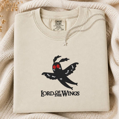 A folded cream-colored crewneck sweatshirt with a black moth graphic and 'LORD OF THE WINGS' text on the front, styled on a beige knit blanket with a necklace draped over the collar. The inner neck label shows the brand 'COMFORT COLORS'.