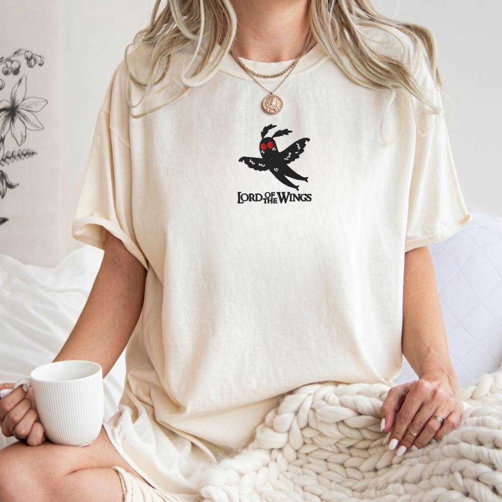 A woman wearing an oversized cream-colored t-shirt with a black 'Lord of the wings' winged logo on the chest, sitting on a bed with a white mug and a chunky knit blanket.
