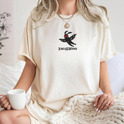 A woman wearing an oversized cream-colored t-shirt with a black 'Lord of the wings' winged logo on the chest, sitting on a bed with a white mug and a chunky knit blanket.