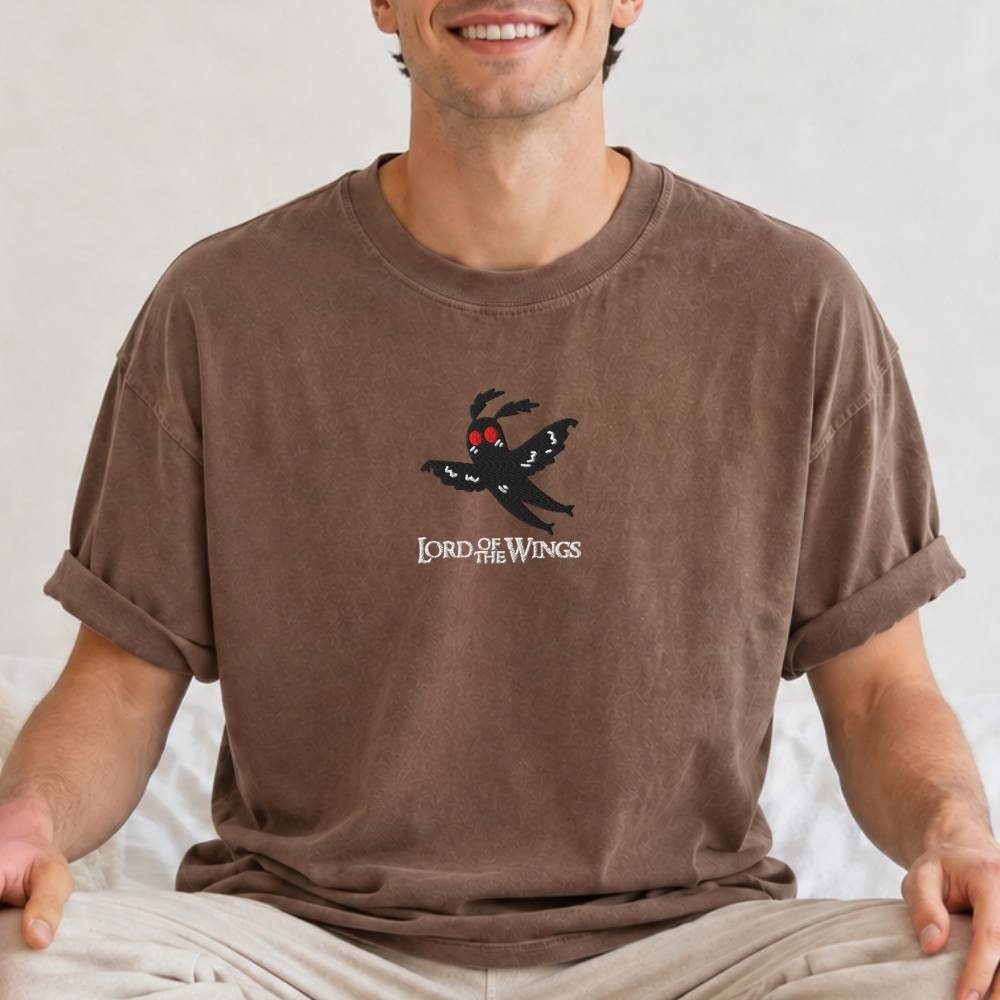 A man wearing a brown t-shirt with a graphic of a black spider with red eyes and the text 'LORD OF THE WINGS' on the chest, sitting against a light background.