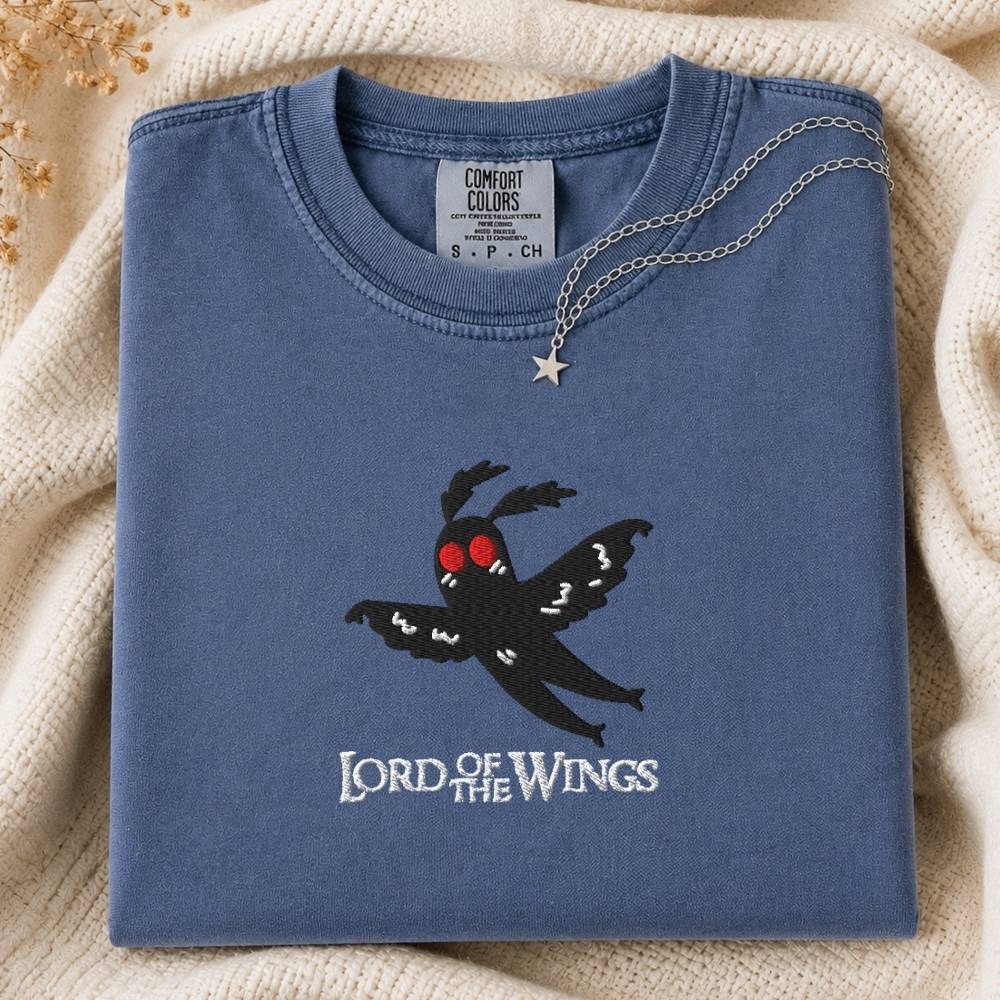 A folded slate blue t-shirt with a black moth graphic and 'LORD OF THE WINGS' text on the front, styled on a cream knit blanket with a silver necklace draped over the collar.