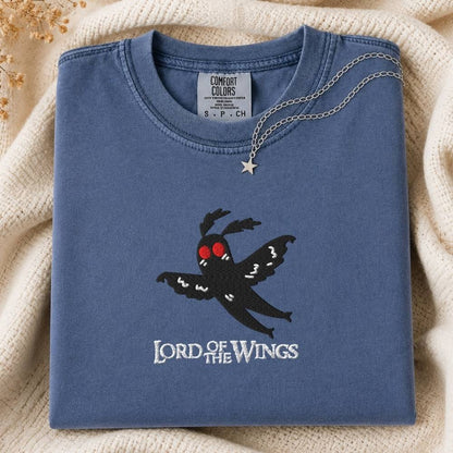 A folded slate blue t-shirt with a black moth graphic and 'LORD OF THE WINGS' text on the front, styled on a cream knit blanket with a silver necklace draped over the collar.