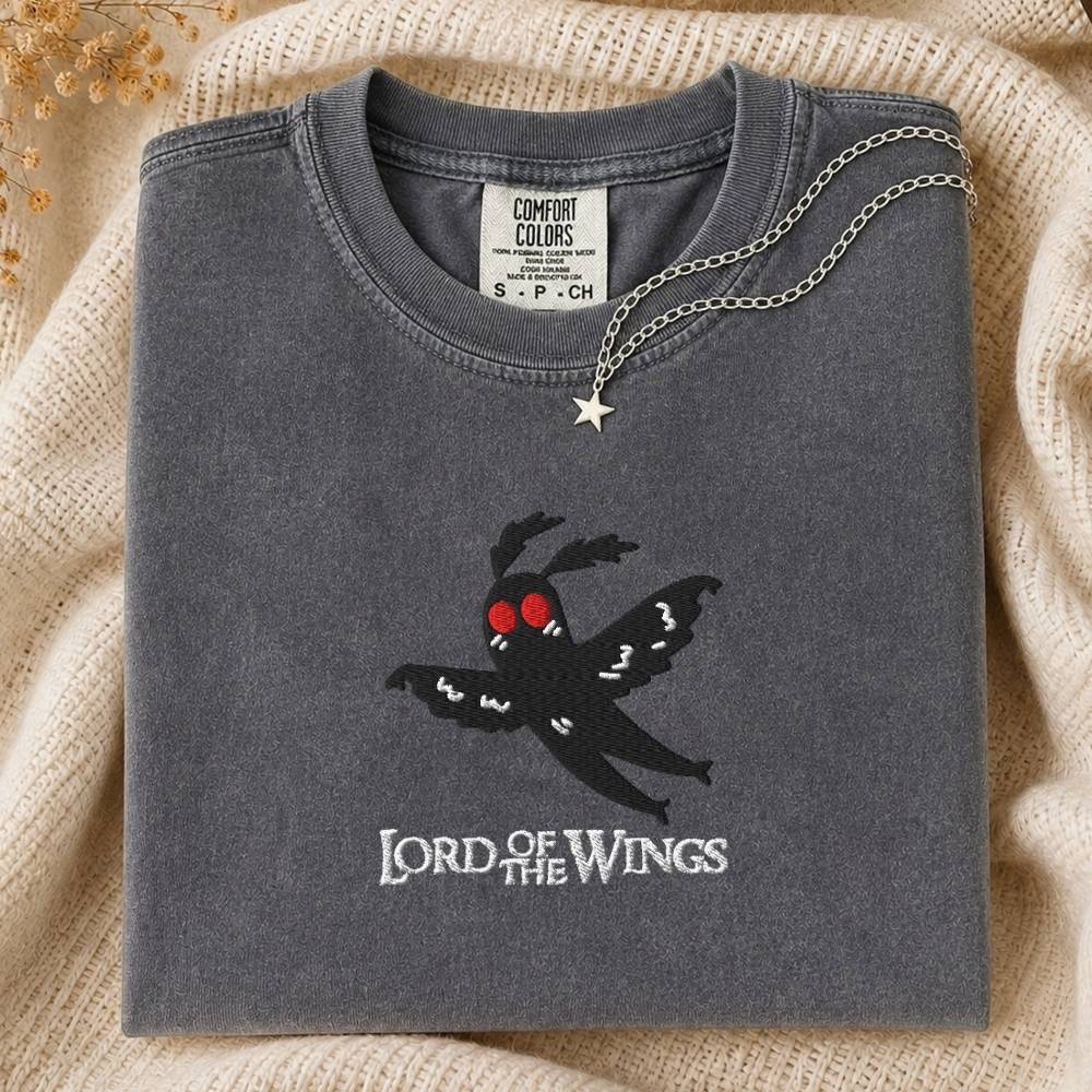 A folded charcoal gray Comfort Colors t-shirt featuring a black embroidered moth with red eyes and the text 'LORD OF THE WINGS' on the front, styled on a beige knit blanket with a silver necklace.