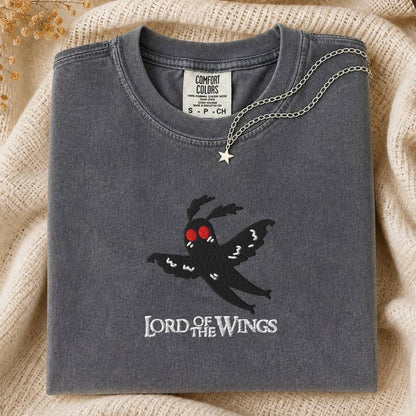 A folded charcoal gray Comfort Colors t-shirt featuring a black embroidered moth with red eyes and the text 'LORD OF THE WINGS' on the front, styled on a beige knit blanket with a silver necklace.