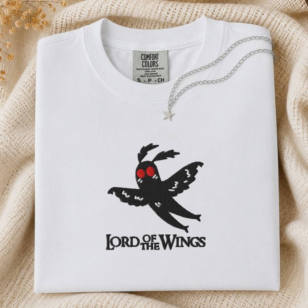 A folded white Comfort Colors t-shirt featuring a black embroidered bird graphic with red eyes and the text 'LORD OF THE WINGS' below it. The shirt is styled on a beige knit blanket with a silver necklace draped over the collar.