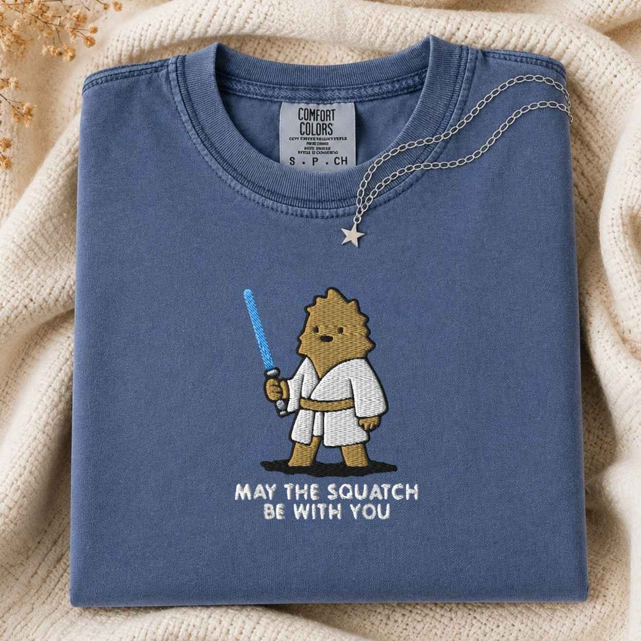 A folded blue Comfort Colors t-shirt with a cartoon squatch character holding a blue laser and the text 'MAY THE SQUATCH BE WITH YOU' underneath. A necklace with a star pendant is draped over the collar, set against a cozy cream-colored knit blanket.