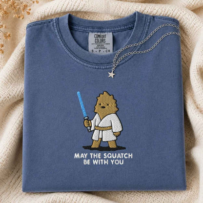 A folded blue Comfort Colors t-shirt with a cartoon squatch character holding a blue laser and the text 'MAY THE SQUATCH BE WITH YOU' underneath. A necklace with a star pendant is draped over the collar, set against a cozy cream-colored knit blanket.