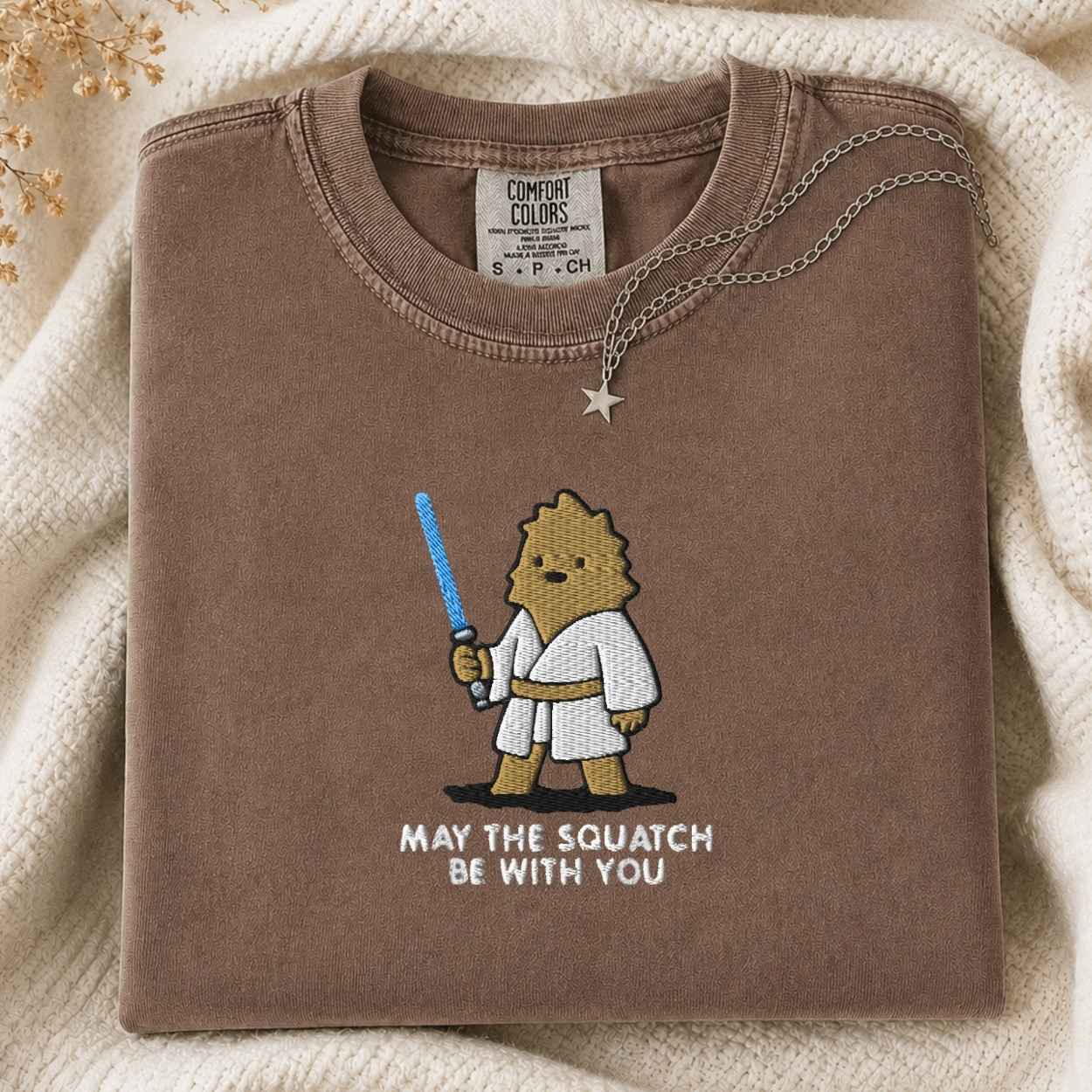 A folded brown Comfort Colors t-shirt with a cartoon squatch character holding a blue laser and the text 'MAY THE SQUATCH BE WITH YOU' underneath. A necklace with a star pendant is draped over the collar, set against a cozy cream-colored knit blanket.