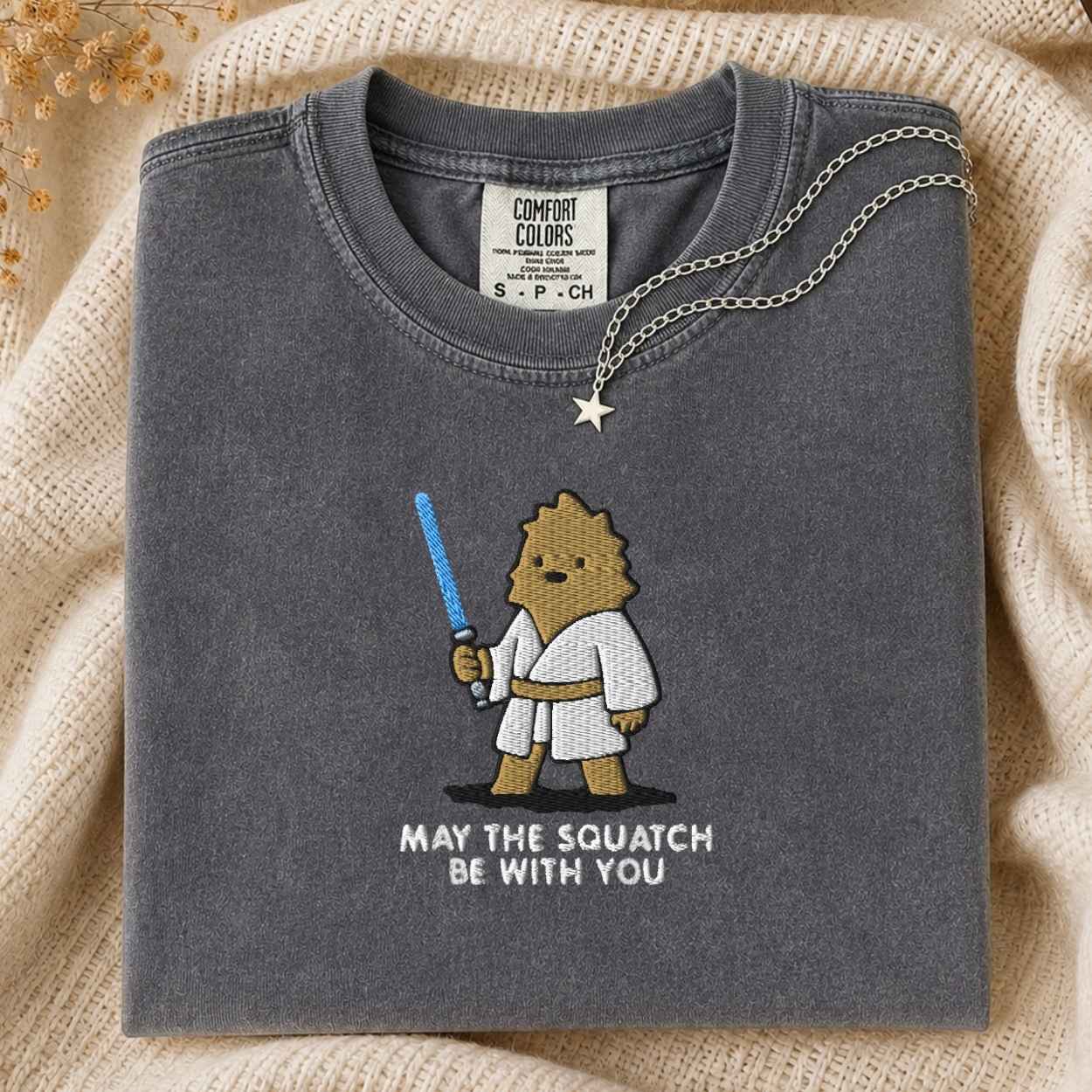 A folded dark gray Comfort Colors t-shirt with a cute cartoon squatch character wearing a white  robe and holding a blue laser sword, with the text 'MAY THE SQUATCH BE WITH YOU' underneath. A silver star pendant necklace is draped over the collar.