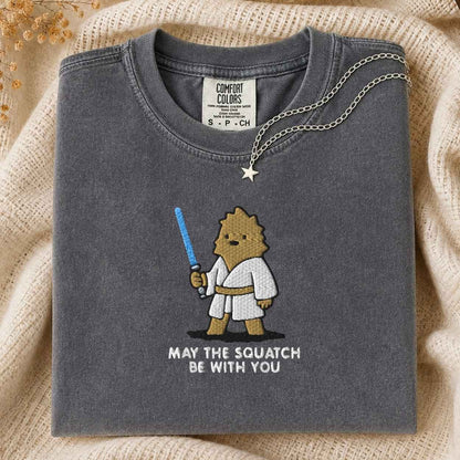 A folded dark gray Comfort Colors t-shirt with a cute cartoon squatch character wearing a white  robe and holding a blue laser sword, with the text 'MAY THE SQUATCH BE WITH YOU' underneath. A silver star pendant necklace is draped over the collar.