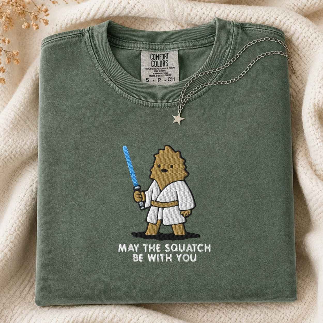 A folded green Comfort Colors t-shirt with a cartoon squatch character holding a blue laser and the text 'MAY THE SQUATCH BE WITH YOU' underneath. A necklace with a star pendant is draped over the collar, set against a cozy cream-colored knit blanket.