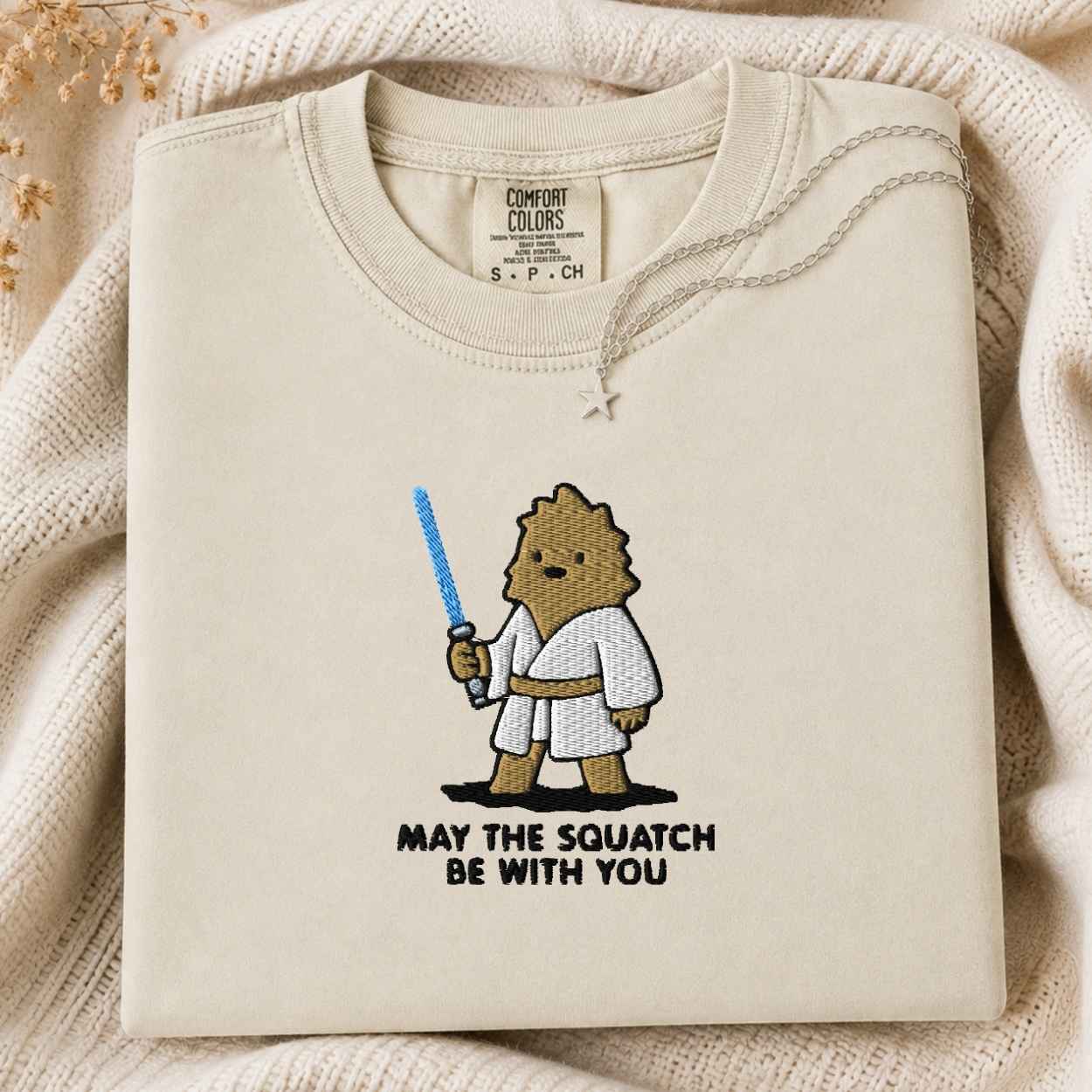 A folded cream-colored tshirt with a cartoon graphic of a squatch character in a white robe holding a blue lightsaber, with the text 'MAY THE SQUATCH BE WITH YOU' underneath. The tshirt is styled on a cozy beige knit blanket with dried flowers in the corner.
