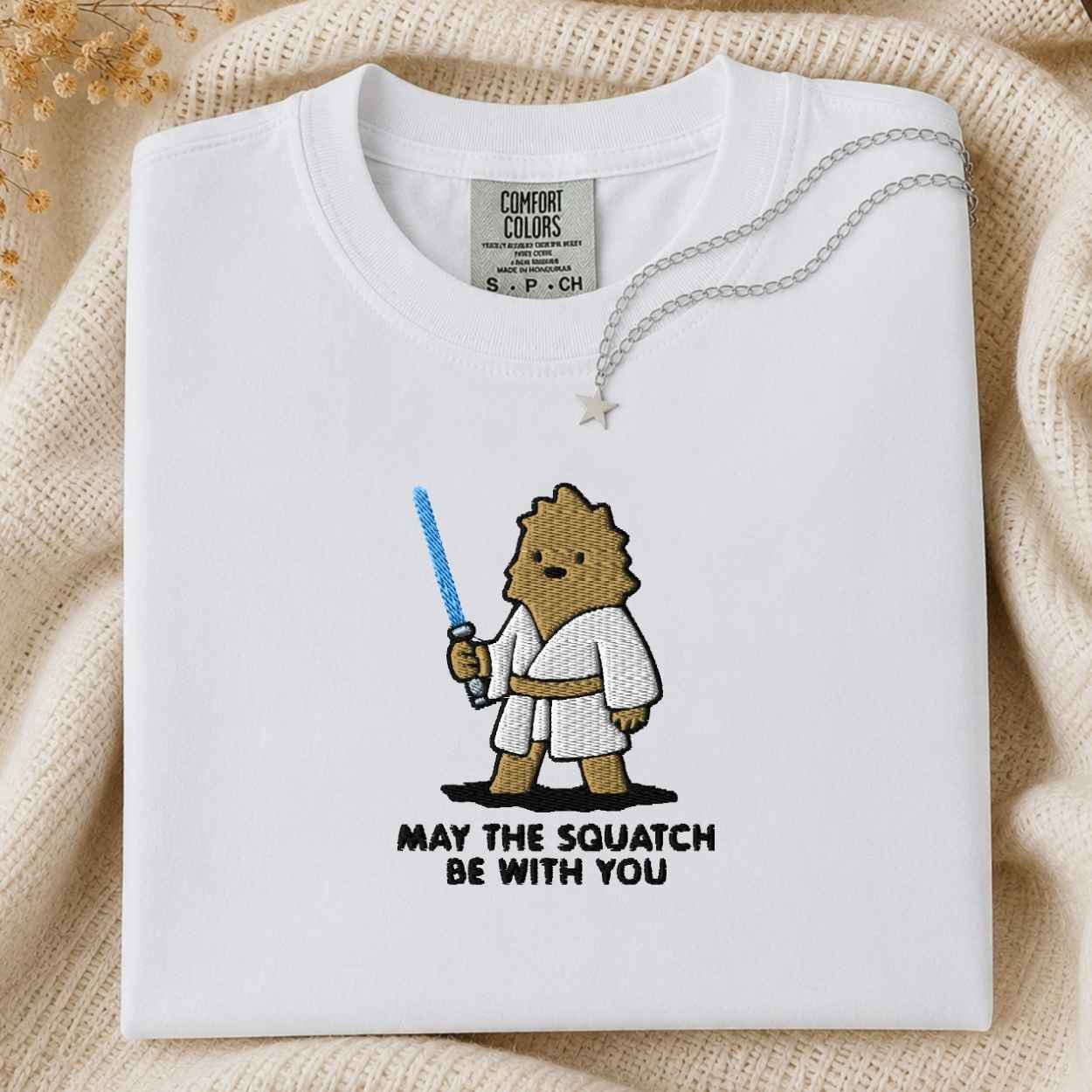 A folded white Comfort Colors t-shirt with a cute embroidered cartoon squatch character holding a blue laser , with the text 'MAY THE SQUATCH BE WITH YOU' below. The shirt is styled on a beige knit blanket with a silver necklace draped over the collar.