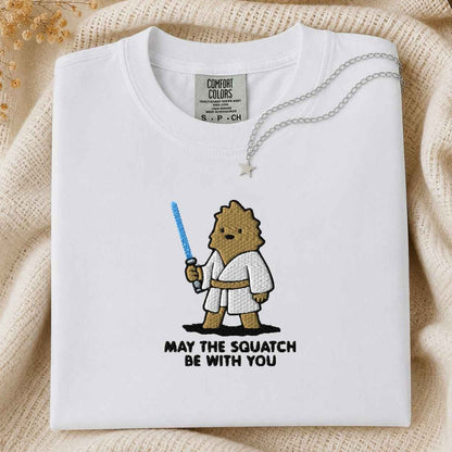 A folded white Comfort Colors t-shirt with a cute embroidered cartoon squatch character holding a blue laser , with the text 'MAY THE SQUATCH BE WITH YOU' below. The shirt is styled on a beige knit blanket with a silver necklace draped over the collar.